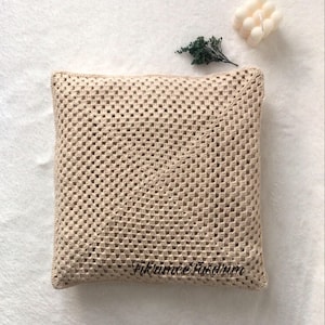 Puede incluir: Una almohada de crochet beige con un patr&oacute;n cuadrado. La almohada est&aacute; sobre una superficie blanca con una peque&ntilde;a planta verde y una vela blanca en el fondo. El texto "Fikrumme Fasarum" es visible en la almohada.