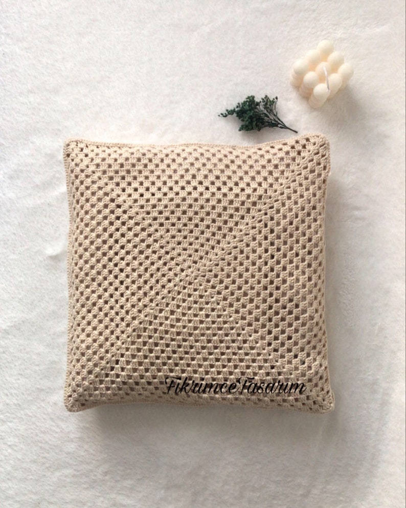 Puede incluir: Una almohada de crochet beige con un patr&oacute;n cuadrado. La almohada est&aacute; sobre una superficie blanca con una peque&ntilde;a planta verde y una vela blanca en el fondo. El texto "Fikrumme Fasarum" es visible en la almohada.