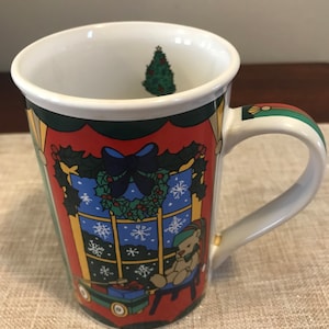 Puede incluir: Una taza de Navidad de cerámica con interior blanco y diseño exterior colorido. El exterior presenta una escena de ventana con un oso de peluche, un árbol de Navidad, copos de nieve y regalos envueltos. El asa es blanca con detalles verdes y dorados.