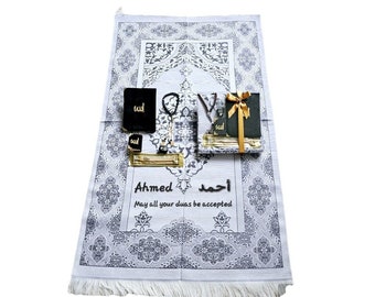 May include: A white prayer rug with a gray floral pattern, featuring a black book, prayer beads, and a gift box tied with a gold ribbon. The rug has the name "Ahmed" and a message printed on it.