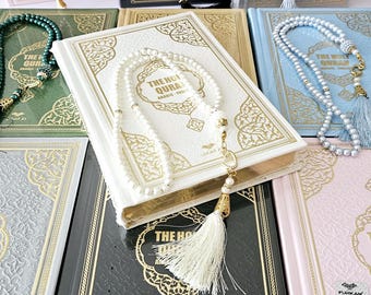 May include: A collection of The Holy Quran books with matching prayer beads in various colors, including pink, black, white, green, blue, and gray. Each book features gold accents and intricate designs.