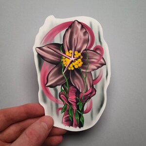 May include: A sticker featuring a detailed illustration of a purple flower with a yellow center and green vines. The flower is surrounded by pink swirls and a white background.