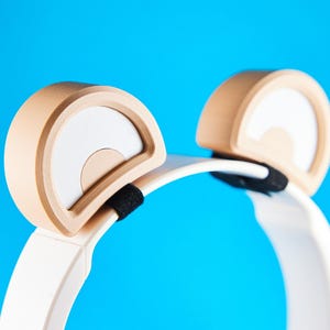 Könnte beinhalten: Kopfhörer mit einem weißen Bügel und beigefarbenen Ohrmuscheln. Jede Ohrmuschel hat ein weißes Halbkreis-Design. Ein schwarzes Stoffstück verbindet die Ohrmuscheln mit dem Bügel. Der Hintergrund ist einfarbig blau.