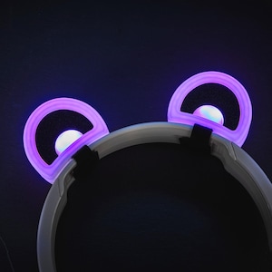 Könnte beinhalten: Nahaufnahme eines Stirnbandes mit zwei leuchtenden, lila Bärenohren. Die Ohren haben einen weißen Innenkreis und einen schwarzen Innenraum. Das Stirnband ist weiß und schwarz, vor dunklem Hintergrund.