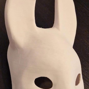 Puede incluir: Una máscara de conejo blanca con dos orejas grandes y agujeros para los ojos. La máscara está hecha de un material blanco liso.