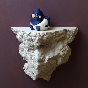 Può includere: Una scultura artigianale di un pinguino con testa blu e corpo bianco, posizionata su una mensola strutturata bianco sporco. La mensola ha un aspetto ruvido, simile alla roccia. Lo sfondo è viola scuro.