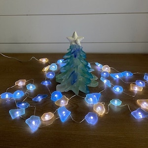 May include: A blue and white glass Christmas tree with a white starfish on top, surrounded by a string of lights with blue, white, and yellow glass bulbs.