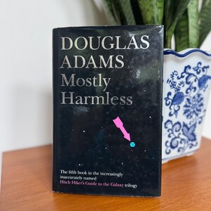 Könnte beinhalten: Ein Buch von Douglas Adams, "Mostly Harmless", mit silbernem Text auf schwarzem Einband. Das Cover-Design zeigt einen rosa Pfeil und einen blauen Punkt. Der Text darunter lautet: "Das fünfte Buch der zunehmend ungenau benannten Hitch Hiker's Guide to the Galaxy Trilogie."