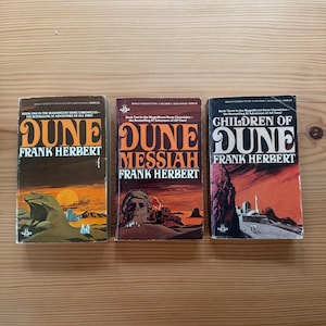 May include: Three vintage paperback books by Frank Herbert from the Dune series. The covers feature illustrations in shades of orange, brown, and green. Titles include "Dune," "Dune Messiah," and "Children of Dune."