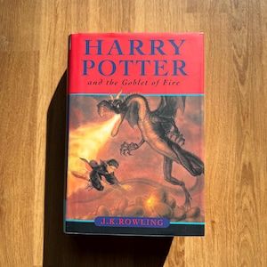 Puede incluir: Un libro de tapa dura titulado "HARRY POTTER and the Goblet of Fire" de J.K. Rowling. La portada presenta un dragón que lanza fuego a una figura, con un fondo rojo y detalles azules. El libro está sobre una superficie de madera.