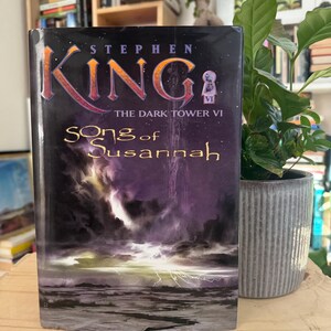 Könnte beinhalten: Ein Taschenbuch mit dem Titel "Song of Susannah" von Stephen King. Es ist das sechste Buch der Reihe "Der Dunkle Turm". Das Cover zeigt einen stürmischen Himmel mit Blitzen und eine dunkle, felsige Landschaft.