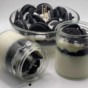 Op de afbeelding: Drie glazen potkaarsen en een glazen komkaars, allemaal ontworpen om op Oreo-koekjes te lijken. De kaarsen hebben lagen witte en zwarte was, met stukjes Oreo-koekjes erop. De komkaars heeft verschillende brandende lonten.