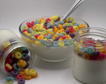 May include: A clear glass bowl and two jars filled with colorful, ring-shaped cereal and white liquid. One jar is tipped over, spilling cereal. A silver spoon rests in the bowl. The cereal pieces are various colors, including red, yellow, green, and purple.