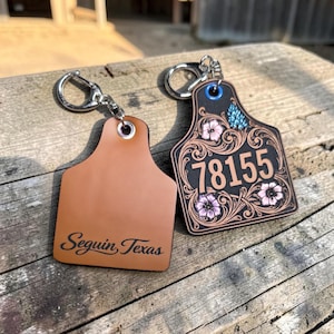 May include: Two leather keychains. One is plain brown with the text "Seguin, Texas". The other is black with the number "78155" and floral designs in pink and blue. Both have silver key rings.