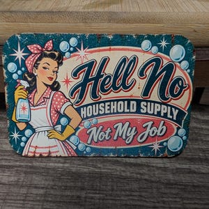 May include: A rectangular metal sign with rounded corners featuring a retro-style illustration of a person in a polka-dot dress and apron holding a spray bottle. The text reads Hell No HOUSEHOLD SUPPLY Not My Job with blue bubbles.