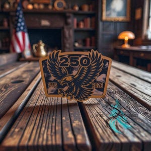 May include: A wooden plaque with a black eagle design and the number "250" above it. The years "1776" and "2026" are on either side of the eagle. The plaque is on a wooden table.
