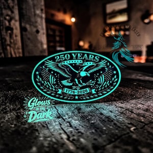 May include: An oval-shaped, glowing-in-the-dark emblem with a patriotic theme. The design features an eagle with outstretched wings, the text "250 YEARS" above, and "1776-2026" below. American flags flank the eagle. The words "Glows in the Dark" are also visible.
