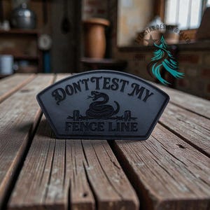 May include: A black, trapezoidal-shaped patch featuring an embossed graphic of a coiled snake positioned above barbed wire. The text "DON'T TEST MY" is arched above the snake, and the words "FENCE LINE" are printed in a straight line below it.