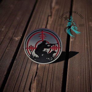 May include: A circular metal emblem featuring a silhouette of a hunter aiming a rifle, with a dog, inside a red and silver target reticle. The emblem is on a wooden surface. The text "EVERSTAR DESIGN & LASER" and "SPIRIT ROOTS" is also visible.