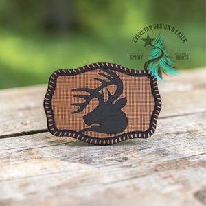 May include: A rectangular patch with rounded, scalloped edges featuring a tan, textured background and a black silhouette of a deer head with large antlers. The patch has a thick black border with a repeating stitch pattern around the perimeter.