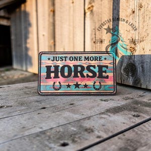 May include: Rectangular sign with the text "JUST ONE MORE HORSE" in black letters. The sign has a turquoise, pink, and brown southwestern design with horseshoes and stars. The sign is framed with a rope border.