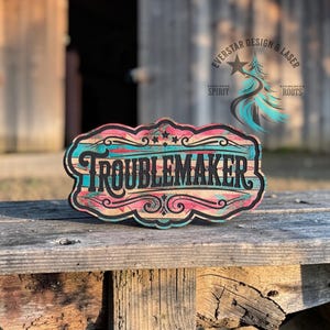 May include: Wooden sign with the word "TROUBLEMAKER" in black script. The sign has a turquoise, pink, and black design with a rustic, distressed look. The sign is on a wooden surface.