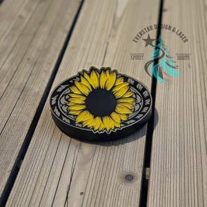 May include: A black and yellow sunflower design belt buckle with an ornate border. The buckle has a black center with a yellow sunflower and is set against a wooden background. The text "EVERSTAR DESIGN & LASER" is visible.