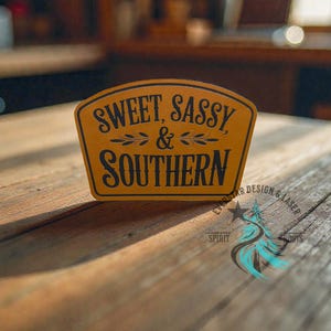 May include: A tan, arched leather patch featuring the black engraved text "SWEET, SASSY, & SOUTHERN" with decorative leaf accents. The patch has a smooth, matte finish and a distinct border, showcasing a clean, professional laser-engraved design.