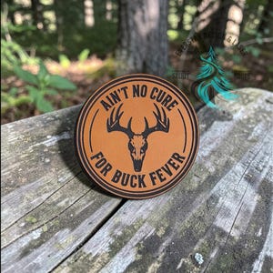 May include: A circular tan leather patch featuring a black engraved deer skull graphic in the center. The text "AIN'T NO CURE FOR BUCK FEVER" is engraved in a circular pattern around the skull, framed by two thin black circular border lines.