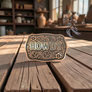 May include: A rectangular, copper-colored belt buckle with the word "HOWDY" in turquoise-colored letters. The buckle has a black background with floral and scrollwork designs. The buckle is sitting on a wooden surface.
