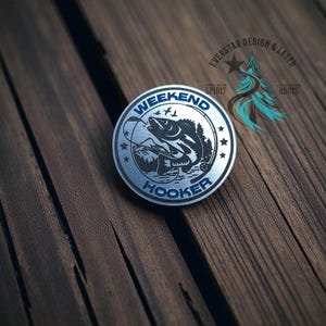 May include: A round, silver-colored metal emblem with the words "WEEKEND HOOKER" in blue lettering. The emblem features a detailed illustration of a fish jumping out of the water, with fishing line and birds. The emblem is set against a wooden background.