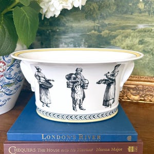 May include: A white ceramic bowl with two side handles, featuring black ink-style illustrations of three figures carrying baskets. The rim and base are accented with a yellow band and a black leaf pattern. The bowl sits atop two stacked books.