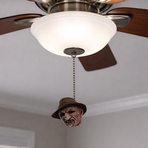 May include: A ceiling fan with wooden blades and a frosted glass light fixture. A Freddy Krueger head hangs from the light fixture's pull chain. The head is wearing a brown hat and has a scarred face.