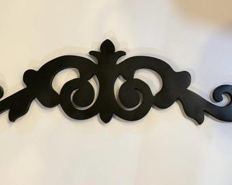 May include: Black decorative wall art with an ornate, symmetrical design. The piece features swirling, scroll-like shapes and a central floral element. This wooden accent adds a touch of elegance to any space.