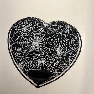 May include: Heart-shaped decorative tray with a black and white spiderweb pattern. The tray has a black trim and a small, round, black stand. A spooky, Halloween-themed accessory.