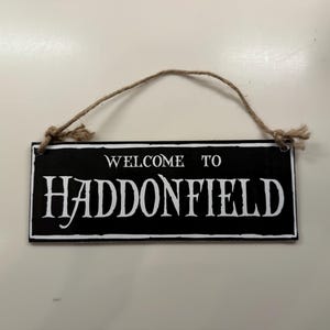 Könnte beinhalten: Ein rechteckiges schwarzes Schild mit weißer Aufschrift "WELCOME TO HADDONFIELD". Es ist mit einer natürlichen Schnur aufgehängt. Ein dekoratives Schild für das Zuhause.