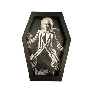 May include: Black and white framed print of a character in a striped suit, set within a coffin-shaped frame. The image features a person with wild, light-colored hair and a dark tie, striking a pose. The frame is a matte black.