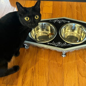 May include: A black cat looking at a black and silver pet food bowl with two stainless steel bowls. The bowl is shaped like a coffin and has decorative silver trim.