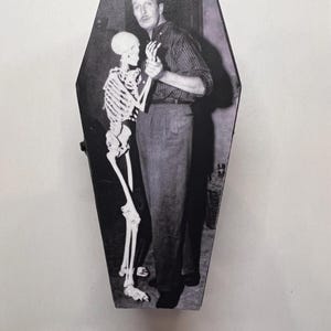 May include: A black and white photo of a man in a striped shirt standing next to a skeleton. The photo is printed on a coffin-shaped box.