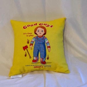 Può includere: Cuscino quadrato giallo con una bambola dei cartoni animati in tuta blu, scarpe rosse e maniche a righe. Il testo "Good Guys" e "It's time to play" è stampato sopra la bambola. Sono raffigurati anche un'ascia e gocce di sangue.