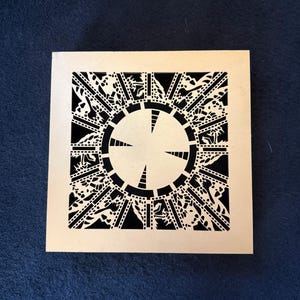 May include: A square, light-colored puzzle box with a detailed black design. The design features a central circular pattern with radiating lines and intricate, symmetrical shapes around the edges. The box is set against a dark blue background.