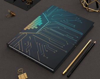 May include: A black notebook with a circuit board design in gold and blue. The notebook is lying on a gray surface with two pencils and a few paper clips.