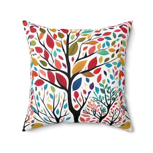 May include: Square cushion featuring a vibrant tree design. The tree has a black trunk and branches adorned with leaves in red, orange, yellow, green, blue, and purple against a white backdrop. The cushion is square.