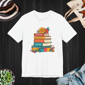 Può includere: T-shirt bianca con un design colorato di libri impilati e fiori. I libri riportano le frasi "IT'S OKAY", "ASK FOR HELP", "MAKE MISTAKES" e "RETRY". Il design include fiori gialli, rossi e rosa.