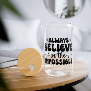 Puede incluir: Un vaso transparente en forma de lata con las palabras "ALWAYS BELIEVE in the IMPOSSIBLE" en negro. Una tapa de madera y una pajita de cristal están junto al vaso. El vaso está sobre una mesa de madera.