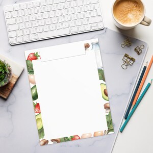 May include: A rectangular notepad with a white center and a decorative border featuring watercolor illustrations of strawberries, avocado halves, brown mushrooms, eggs, and a glass milk bottle, arranged on a white surface with office supplies.