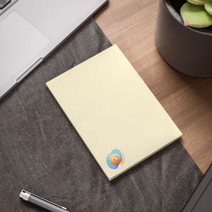 Könnte beinhalten: Ein cremefarbener Haftnotizblock mit einer bunten Cartoon-Grafik in der Ecke. Die Notizen liegen auf einem Holzschreibtisch neben einem Laptop und einem Stift. Eine Topfpflanze befindet sich im Hintergrund.