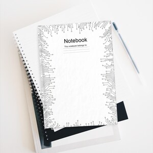 May include: White spiral-bound notebook with a black circuit board design border. The cover reads "Notebook" and "This notebook belongs to:" with a blank line. A pen rests on the white surface next to the notebook.