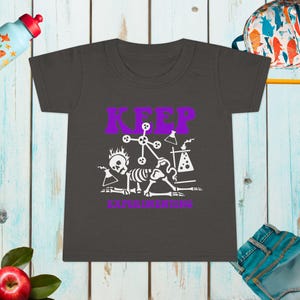 Könnte beinhalten: Dunkelgraues Kleinkind-T-Shirt mit einem lila-weißen Aufdruck. Der Aufdruck zeigt das Wort "KEEP" über "EXPERIMENTING", zusammen mit Skelettfiguren und wissenschaftlichen Illustrationen. Eine Trinkflasche und ein Rucksack sind im Hintergrund zu sehen.