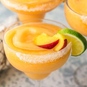 May include: Three frozen peach margaritas in margarita glasses with a sugared rim. Each glass is garnished with a slice of peach and a lime wedge.